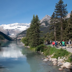 a group of people walking along a river with trees and mountains in the background