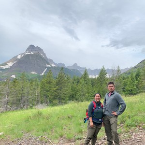 a man and woman standing on a trail with trees and mountains in the background